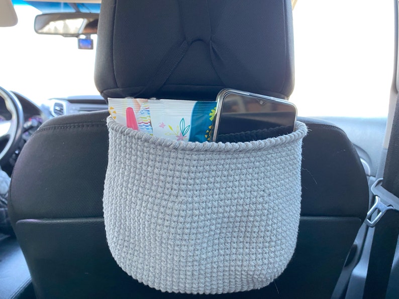 May include: A gray crocheted car seat organizer hanging from the headrest of a car seat. The organizer is filled with a package of wipes and a smartphone.