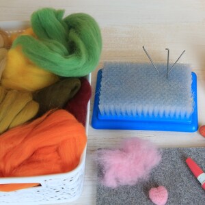 May include: A craft setup featuring a white basket filled with colorful wool roving in shades of green, yellow, orange, and brown. A blue needle felting tool, pink wool, and a gray felt mat are also present, suggesting a fiber art project.