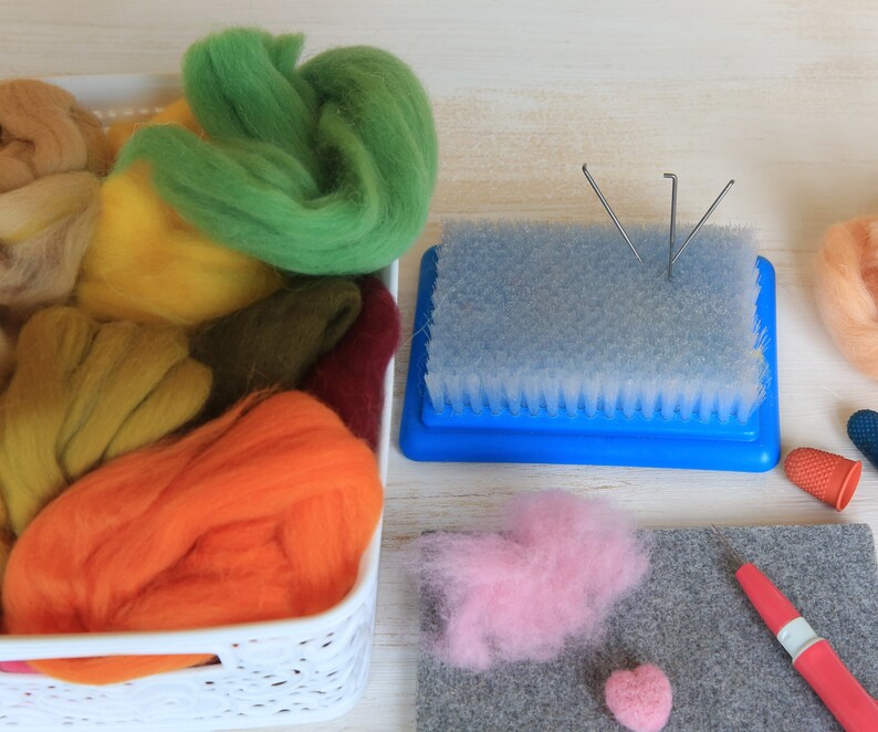 May include: A craft setup featuring a white basket filled with colorful wool roving in shades of green, yellow, orange, and brown. A blue needle felting tool, pink wool, and a gray felt mat are also present, suggesting a fiber art project.
