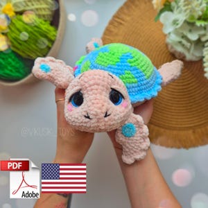 May include: A crocheted turtle amigurumi toy with a pink body, blue and green shell, and large blue eyes. The toy is being held by a person's hands.