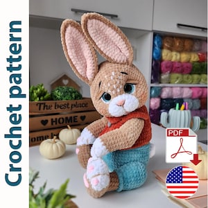 Puede incluir: Patrón de crochet para un conejito marrón y blanco que lleva una camisa roja y pantalones vaqueros azules. El patrón está disponible para descargar en PDF. La imagen también incluye el texto "PDF" y un icono de descarga.