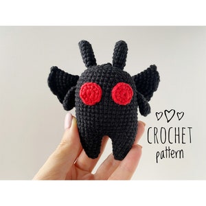 May include: A black crochet creature with red eyes and small wings. The text "CROCHET pattern" is written in black script. The creature is held in a hand against a white background.