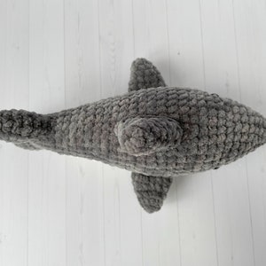 Puede incluir: Un juguete de peluche de tibur&oacute;n gris tejido a crochet. El tibur&oacute;n tiene una superficie texturizada y peluda y una aleta dorsal.