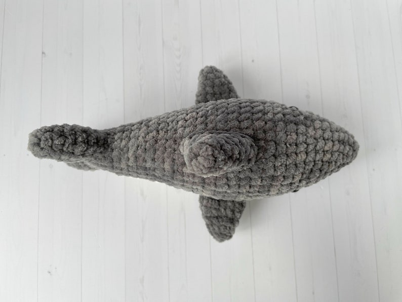 Puede incluir: Un juguete de peluche de tibur&oacute;n gris tejido a crochet. El tibur&oacute;n tiene una superficie texturizada y peluda y una aleta dorsal.