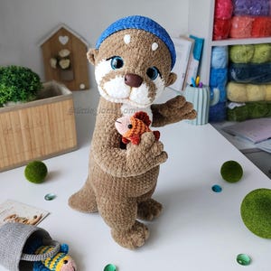 Adorable crochet otter plushie with a blue beanie, hugging a small orange fish. Soft, handmade amigurumi animal pattern for beginner or intermediate crocheters.