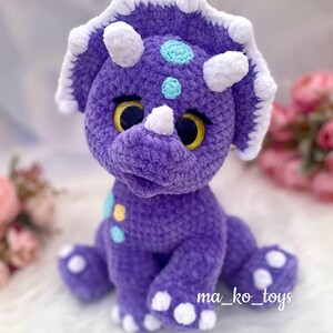 May include: Purple crocheted triceratops stuffed animal with white accents and yellow eyes. The toy has a light blue and yellow polka dot pattern on its body. The text "ma_ko_toys" is visible on the bottom right corner of the image.