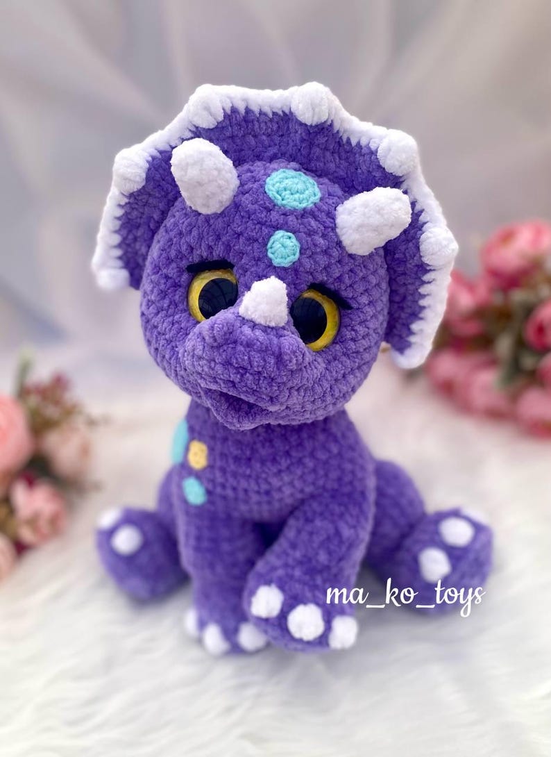 May include: Purple crocheted triceratops stuffed animal with white accents and yellow eyes. The toy has a light blue and yellow polka dot pattern on its body. The text "ma_ko_toys" is visible on the bottom right corner of the image.
