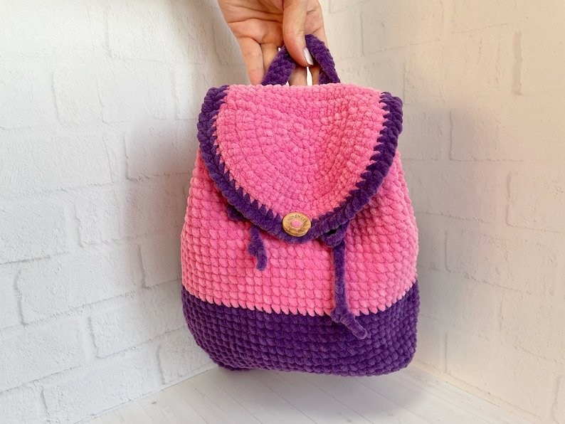 Pu&ograve; includere: Uno zaino in crochet rosa e viola con chiusura a bottone in legno. Lo zaino ha una chiusura a coulisse ed &egrave; realizzato con un filato morbido e soffice.