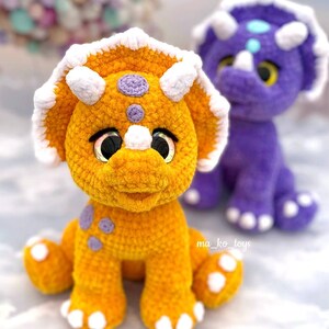 May include: Two crocheted dinosaur toys, one orange and one purple, with white accents and large eyes. The orange dinosaur is sitting on a light gray surface.