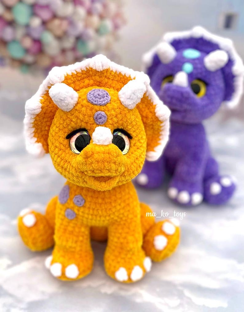 May include: Two crocheted dinosaur toys, one orange and one purple, with white accents and large eyes. The orange dinosaur is sitting on a light gray surface.