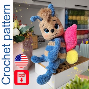 Peut inclure: Poupée abeille au crochet faite à la main, corps rayé bleu et beige, aile rose et queue jaune. La poupée a un visage souriant avec des yeux bleus. L'image comprend le texte "Crochet pattern" et une icône du drapeau américain.
