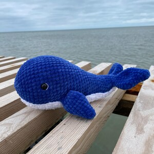 May include: A blue crocheted whale plush toy with white accents and black eyes, sitting on a wooden surface.