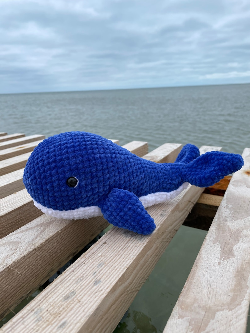 May include: A blue crocheted whale plush toy with white accents and black eyes, sitting on a wooden surface.