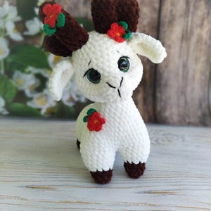 May include: A white crocheted goat with brown yarn antlers and red flower accents. The goat has green eyes and a brown nose. The goat is standing on a wooden surface.
