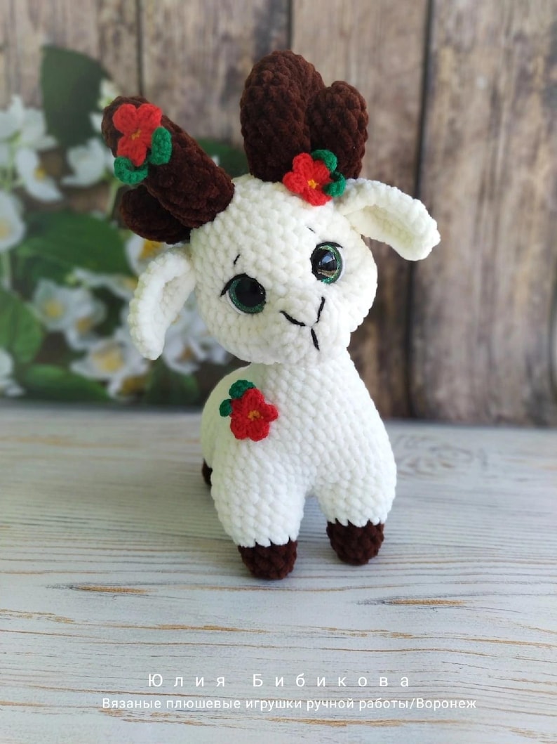 May include: A white crocheted goat with brown yarn antlers and red flower accents. The goat has green eyes and a brown nose. The goat is standing on a wooden surface.