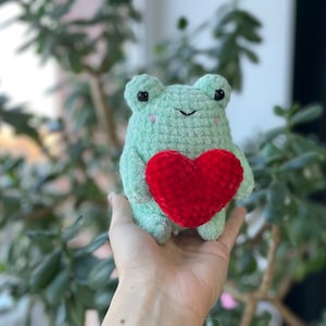 May include: A green crocheted frog holding a red crocheted heart. The frog has black eyes and a small smile.