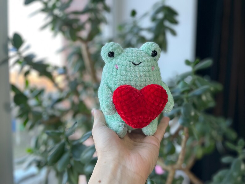 May include: A green crocheted frog holding a red crocheted heart. The frog has black eyes and a small smile.