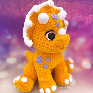 May include: A crocheted yellow triceratops dinosaur toy with white and purple accents. The toy has large, pink eyes and a friendly expression. The toy is sitting on a purple and white background.