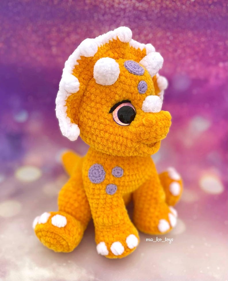 May include: A crocheted yellow triceratops dinosaur toy with white and purple accents. The toy has large, pink eyes and a friendly expression. The toy is sitting on a purple and white background.