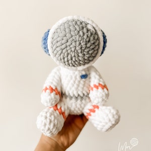 May include: A white crocheted astronaut toy with grey and blue details. The astronaut has a round head with a white helmet and a grey face. The astronaut is wearing a white suit with pink and red stripes.