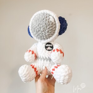 May include: White crocheted astronaut toy with red and blue accents. The astronaut has a grey crocheted helmet and a round patch that says "MNMS".