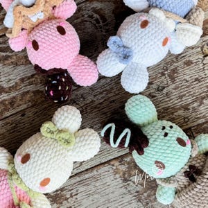 May include: A collection of handmade crochet bunny plushies in various pastel colors. Each bunny features a round head, floppy ears, and a decorative element, such as a bow or a chocolate-covered treat. The plushies are arranged on a wooden surface.