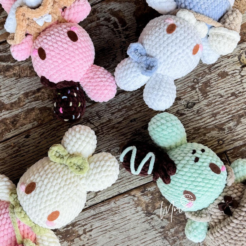 May include: A collection of handmade crochet bunny plushies in various pastel colors. Each bunny features a round head, floppy ears, and a decorative element, such as a bow or a chocolate-covered treat. The plushies are arranged on a wooden surface.