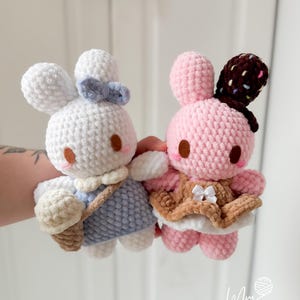 May include: Two handmade crochet bunny dolls. One is white with a blue dress and bow, holding a beige ice cream cone. The other is pink with a brown and white chocolate-covered ear and a brown vest with a white bow.
