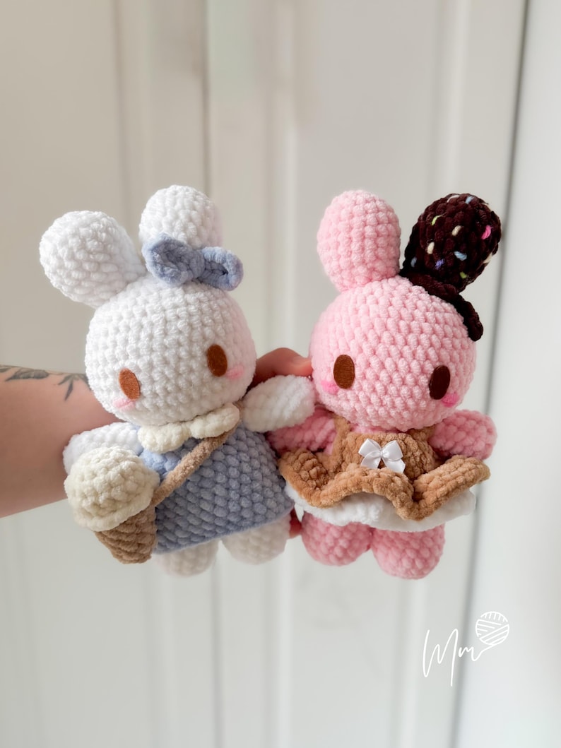 May include: Two handmade crochet bunny dolls. One is white with a blue dress and bow, holding a beige ice cream cone. The other is pink with a brown and white chocolate-covered ear and a brown vest with a white bow.