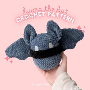 Op de afbeelding: Een gehaakte vleermuisknuffel in blauwtinten, met een zwarte band en witte ogen. De tekst "luma the bat CROCHET PATTERN" staat erboven. De knuffel wordt tegen een roze achtergrond gehouden.