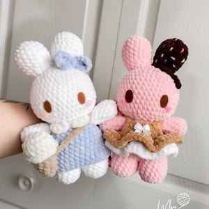 May include: Two handmade crochet bunny dolls. One is white with a blue bow and dress, carrying a beige bag. The other is pink with a brown hat and dress. Both have brown eyes and are made of soft yarn.