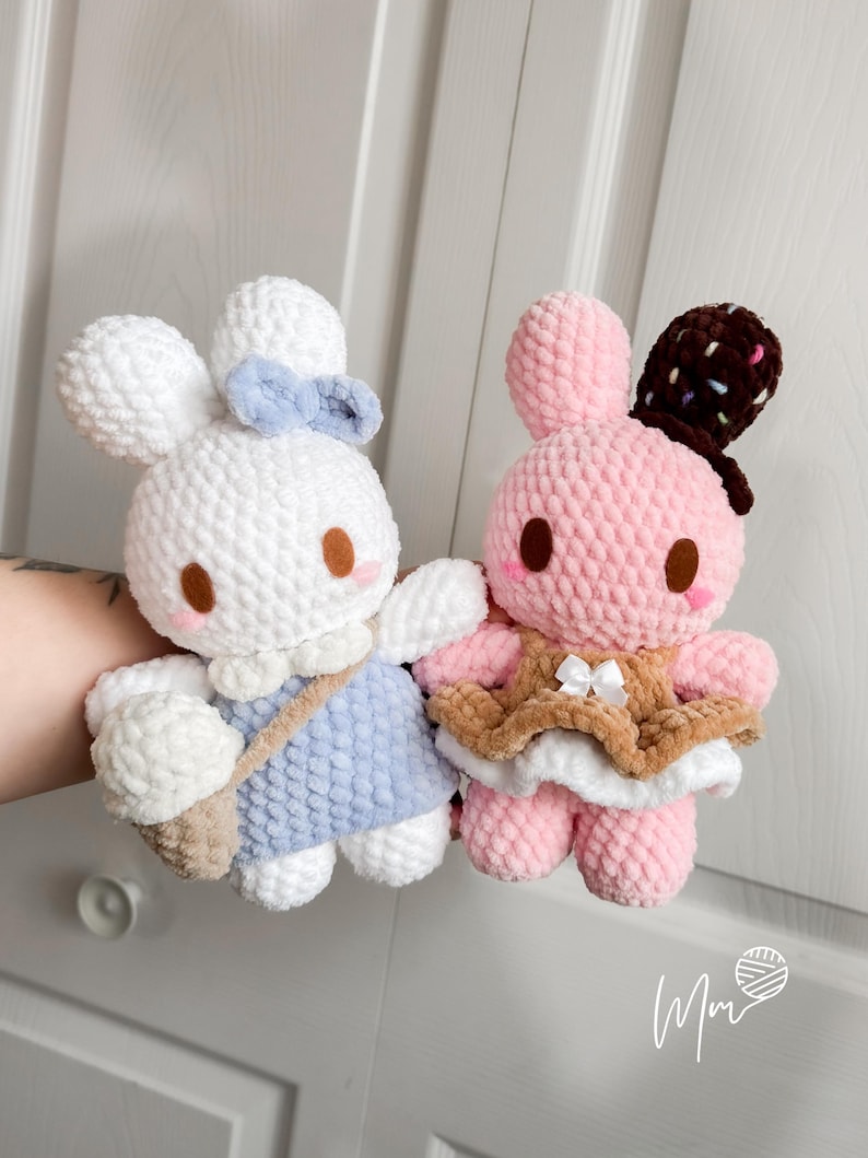 May include: Two handmade crochet bunny dolls. One is white with a blue bow and dress, carrying a beige bag. The other is pink with a brown hat and dress. Both have brown eyes and are made of soft yarn.
