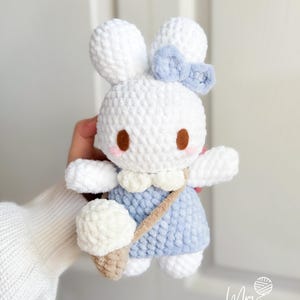 May include: A white crochet bunny doll with a blue dress and bow. The bunny has brown eyes and a beige ice cream cone. The doll has a beige strap across its body. The doll is handmade.
