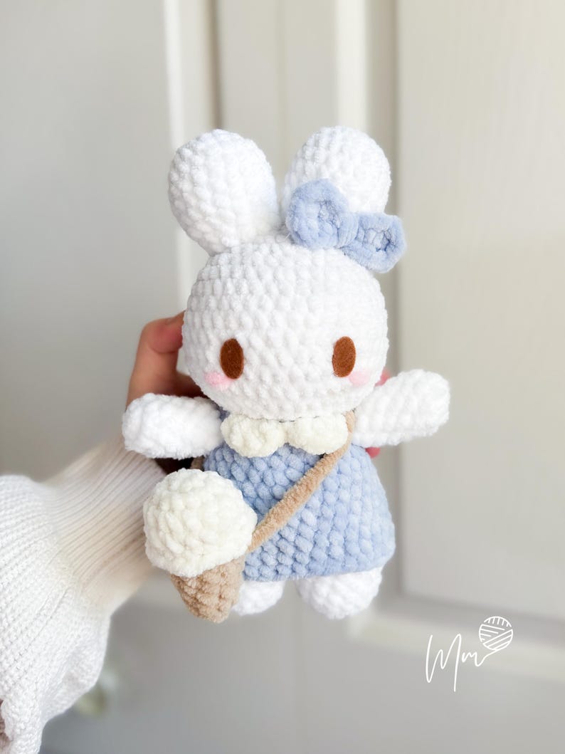 May include: A white crochet bunny doll with a blue dress and bow. The bunny has brown eyes and a beige ice cream cone. The doll has a beige strap across its body. The doll is handmade.