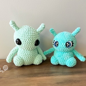 May include: Two crocheted plush toys in the shape of aliens. Both are light green with black eyes. The smaller alien has blue eyes with white stars on them.
