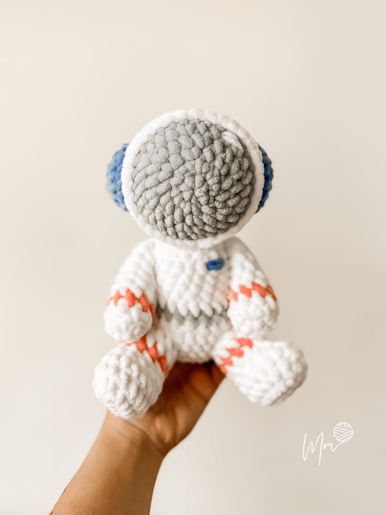 May include: A white crocheted astronaut toy with grey and blue details. The astronaut has a round head with a white helmet and a grey face. The astronaut is wearing a white suit with pink and red stripes.