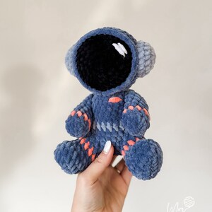 May include: A crocheted blue astronaut toy with a black face and orange accents. The toy is being held by a hand.