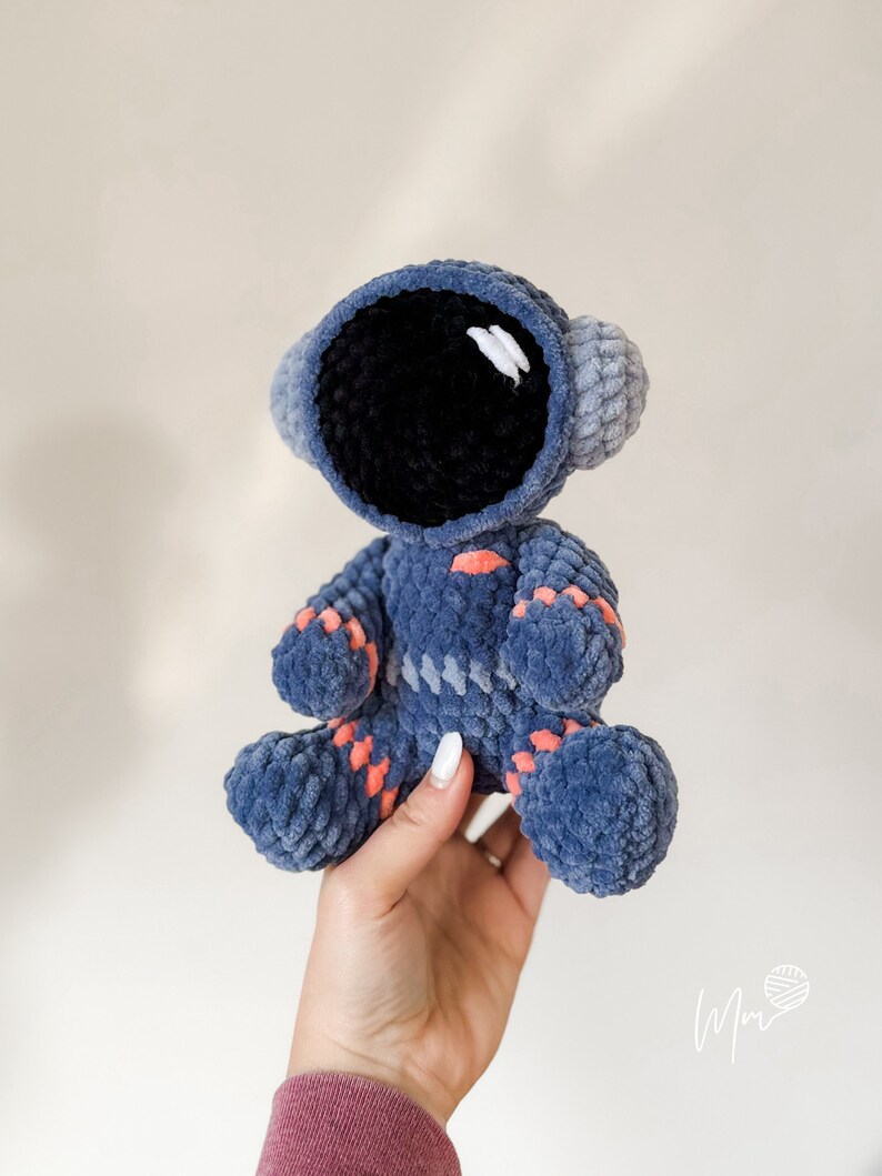 May include: A crocheted blue astronaut toy with a black face and orange accents. The toy is being held by a hand.