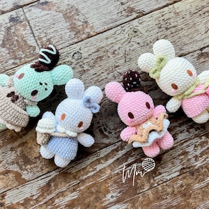 May include: Four handmade crochet bunny dolls in pastel colors, each with unique accessories. One is mint green with a chocolate swirl, another is white with a blue bow, a third is pink with a star, and the last is white with a pink scarf. They are arranged on a wooden surface.