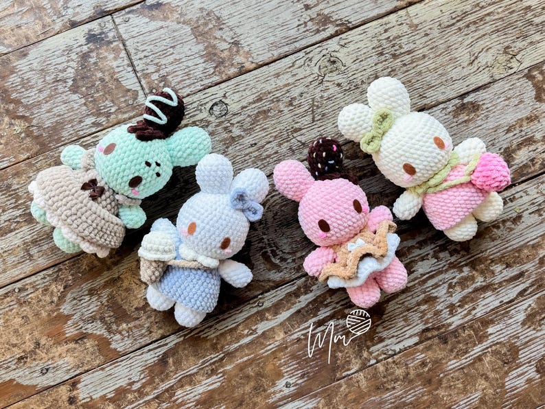 May include: Four handmade crochet bunny dolls in pastel colors, each with unique accessories. One is mint green with a chocolate swirl, another is white with a blue bow, a third is pink with a star, and the last is white with a pink scarf. They are arranged on a wooden surface.