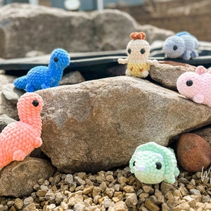May include: A collection of crocheted dinosaur toys in various colors, including pink, blue, green, and white, sitting on a bed of rocks.