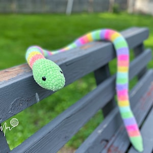 以下が含まれることがあります： 緑色の頭と黒い目をした虹色の毛糸で編まれた蛇のぬいぐるみが、灰色の木のベンチの上に置かれています