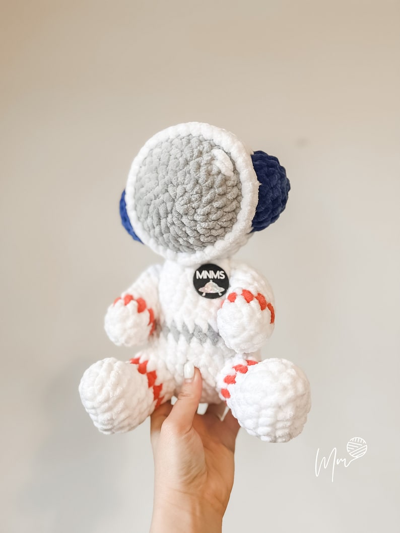 May include: White crocheted astronaut toy with red and blue accents. The astronaut has a grey crocheted helmet and a round patch that says "MNMS".