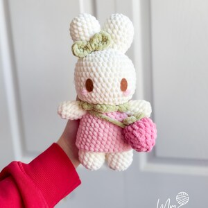 May include: A handmade white crochet bunny doll with a pink dress and a green bow. The bunny has brown eyes and pink cheeks, and carries a pink purse. The doll is soft and plush, perfect for a child's toy or nursery decor.