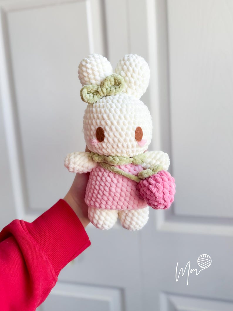 May include: A handmade white crochet bunny doll with a pink dress and a green bow. The bunny has brown eyes and pink cheeks, and carries a pink purse. The doll is soft and plush, perfect for a child's toy or nursery decor.