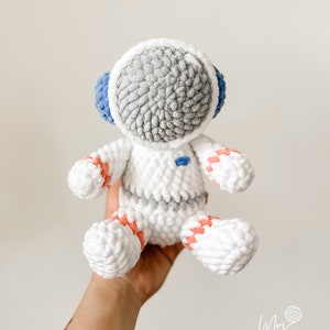 May include: A white crocheted astronaut toy with a grey head, blue visor, and orange and white stripes on the arms and legs.