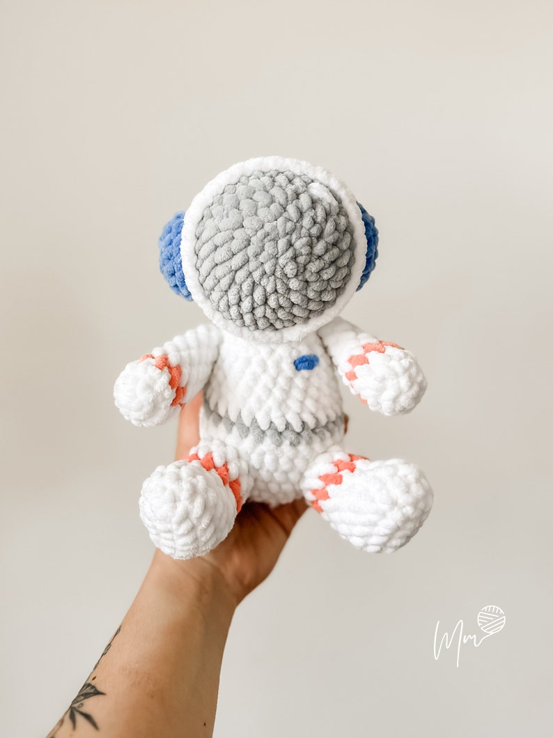 May include: A white crocheted astronaut toy with a grey head, blue visor, and orange and white stripes on the arms and legs.
