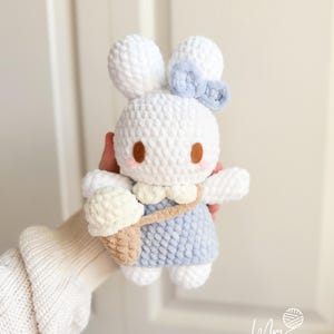 May include: A handmade white crochet bunny doll with a light blue dress and a matching bow. The bunny has brown eyes and pink cheeks. It carries a beige ice cream cone in a small bag. The doll is soft and plush.