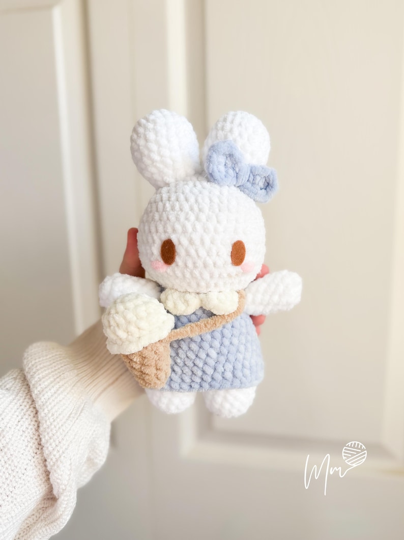 May include: A handmade white crochet bunny doll with a light blue dress and a matching bow. The bunny has brown eyes and pink cheeks. It carries a beige ice cream cone in a small bag. The doll is soft and plush.