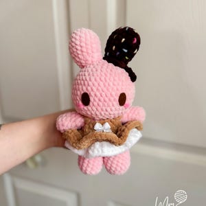 May include: A pink crocheted bunny plushie with brown eyes and a chocolate-covered ear. The bunny wears a brown and white dress with a small white bow. The plush toy is soft and cuddly, perfect for children.